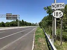 A road sign reading north above a black rectangle with a white oval containing 185 in black numerals. Trees and industrial buildings can be seen in the background.