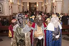 Three men disguised as kings followed by a retinue in a hall.