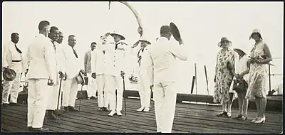 Several men dressed in white tropical uniforms on the deck of a ship, two men wearing pith helmets are saluting, and a group of women wearing dresses and hats are standing to one side