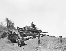 A main battle tank in a hull down position, its turret traversed to aft. In the foreground is a soldier kneeling.