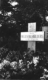 Black-and-white photograph of a wooden cross on a grave, bearing the inscription "Oberst Werner Mölders, 18. 3. 1913 – 22. 11. 1944."