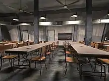 A grey wall with two doors is in the background, a chalkboard in its centre, and ceiling fans and hanging lights are overhead. In the foreground are two long cafeteria tables extending into the background, each having ten chairs; other tables are partially visible to the left and right.