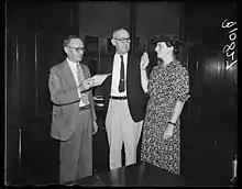 Two white men and a white woman, all standing; the woman has her hand raised in a position of swearing an oath; the woman is wearing a print dress, and the men are wearing suit jackets and ties