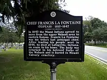 A historic marker with the following text, “CHIEF FRANCIS LA FONTAINE (TOPEAH) 1810-1847; In 1840 the Miami Indians agreed to move from the upper Wabash area to eastern Kansas. Francis La Fontaine was his tribe’s last principal chief. After leading his people west in 1846, he died at Lafayette, Indiana, en route to his home. The body was returned to his beloved Forks of the Wabash and is buried in Mount Calvary cemetery.”