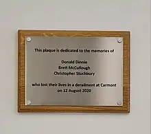 a simple plaque in brushed steel on a wooden backing, which reads "This plaque is dedicated to the memories of Donald Dinnie, Brett McCullough, Christopher Stuchberry who lost their lives in a derailment at Carmont on 12 August 2020"