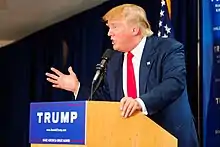 Trump speaking behind a brown wooden podium, wearing a dark blue suit and a red tie. The podium sports a blue "TRUMP" sign.