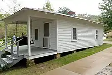 Present-day photograph of a whitewashed house, about 15&nbsp;feet wide. Four bannistered steps in the foreground lead up to a roofed porch that holds a swing wide enough for two. The front of the house has a door and a single-paned window. The visible side of the house, about 30&nbsp;feet long, has double-paned windows.