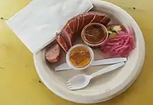 Photograph of sausage and other food items on a paper plate with plastic cutlery and a white paper napkin
