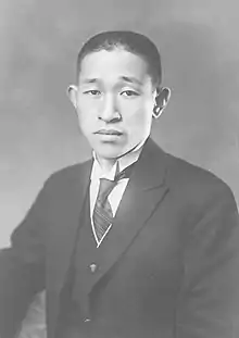 Black and white portrait. Head shot of Matsushita in front of a dark background, wearing a dark suit jacket with a light wing-collared shirt, dark vest, and dark striped necktie.