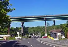 A pair of bridges pass from left to right on tall supports that tower over the other structures in the area, such as telephone poles and single-story buildings. The two-lane NY 22 passes underneath the bridge and proceeds into the background.