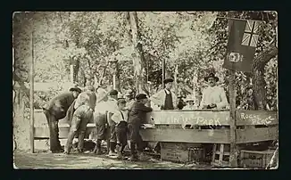 A group of men and young boys are standing at a stall set up in a park at Roche Perce sometime between 1910 and 1925. Writing on the image indicates this is a park in Roche Perce.
