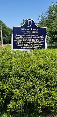 A historical marker surrounded by a bush with small purple flowers. The sign reads, "Indiana School for the Blind. Established 1847 by the General Assembly to provide education for Indiana's blind children. School on downtown site, 1848-1930. Relocated 1930 to present site on North College Avenue. Provides services to blind and visually impaired school-age children.