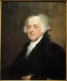 Partially bald man with white hair in black suit sits