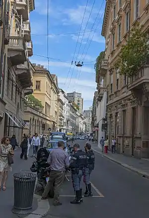 A narrow street slightly curving rightwards in the distance, above which are overhead wires and a blue sky. On either side are low multistory buildings painted in light colors with ornate architectural decoration. In the foreground are several men in police uniforms clustered in front of a motorcycle with a man in plain clothes, their backs to the camera. People are walking along both sides of the street.