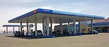 A large gas station with a blue canopy with the words "Manaseer" and the company logo on it. Below it are pumps marked as "diesel" in English and Arabic. In the rear is a large one-story building with "Administration offices" on it