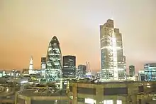 A skyline of modern buildings underneath an orange sky. All the skyscrapers are lit up by office lights from several windows.