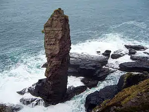 A tall sea stack consisting of a layered sedimentary rock is situated off a rocky coast and sits amid breaking waves and foamy waters.