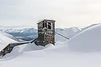 "A wooden building with slanted roofs and a cable coming out of one of the towers. Large, smooth drifts abut the building on one side, and only a three-story tower is fully exposed. In the background is a wide valley, with more mountains on the other side."