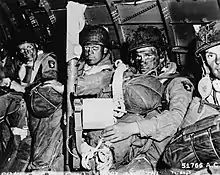 A black and white image of three men and part of a fourth in combat gear, with their faces smeared with black, sitting against a curved metallic wall and carrying heavy packs of gear