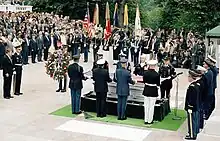 Four men, in military uniforms with different combinations of white and blue, stand on either side of a coffin at lower right, out of doors on a stone floor. Behind them are a group of flags. In the background and at left is an audience on risers