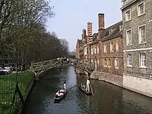 Colour photograph of two punts going under a wooden bridge on the River Cam. On the right of the image there are buildings with foundations built directly into the river, and on the left, a grass riverbank.