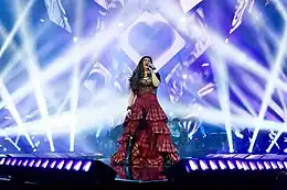 Velasquez sing to a hand held microphone wearing a tartan skirt and black bodysuit