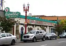 Photograph of the exterior of a building with a neon sign, parking pay station, and red lamp post; cars line the street.