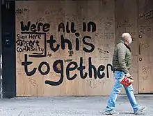 A photo of a boarded-up business in New York during the COVID-19 pandemic, featuring a masked bald man in jeans and a coat passing by; graffiti text on the boarding reads "We're ALL in this together"