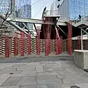 This image shows an outdoor urban installation consisting of multiple thin red rods arranged in clusters, creating a striking visual contrast against the gray paving stones, a brick wall, and the metal framework of an overhead pedestrian bridge.