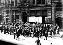 Black and white image of building exterior, including a group of people reading news bulletin boards