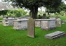 Three-quarter view of several large stone tombs and some gravestones in a grassed churchyard, with trees visible in the background.
