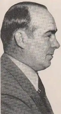 Black and White picture. Profile of a man facing right. He has tidy hairs and wears a suit with a cravat. We can only see the bust.