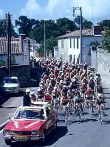 A road with, from front to back, a motor, a car, and tens of cyclists