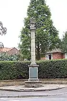 tall, thin gothic revival memorial. A tall round pillar on a square base in a residential area, a bungalow discretely behind a hedge. Behind the pillar is a neatly clipped hedge and a tall, green conifer.