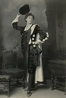  Young woman with short curly hair standing in flamboyant male clothing, with cape in indoor setting