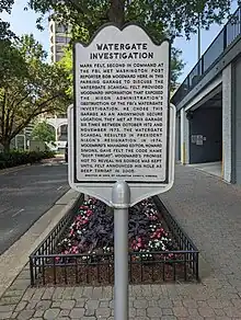 Photo of a grey historical marker titled "Watergate Investigation" on the sidewalk beside a small urban street with a parking garage door visible on the right behind the sign. The marker reads: "Mark Felt, second in command at the FBI, met Washington Post reporter Bob Woodward here in this parking garage to discuss the Watergate scandal. Felt provided Woodward information that expose the Nixon Administration’s obstruction of the FBI’s Watergate investigation. He chose this garage as an anonymous secure location. They met at this garage six times between October 1972 and November 1973. The Watergate scandal resulted in President Nixon’s resignation in 1974. Woodward’s managing editor, Howard Simons, gave Felt the code name ‘Deep Throat’. Woodward’s promise not to reveal his source was kept until Felt announced his role as Deep Throat in 2005. Erected in 2008 by Arlington County, Virginia.”