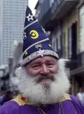 A bearded white man in bright purple and gold robes smiles towards the camera. On his head, a tall pointed purple cap emblazoned with silver and gold stars, moons, and crowns.